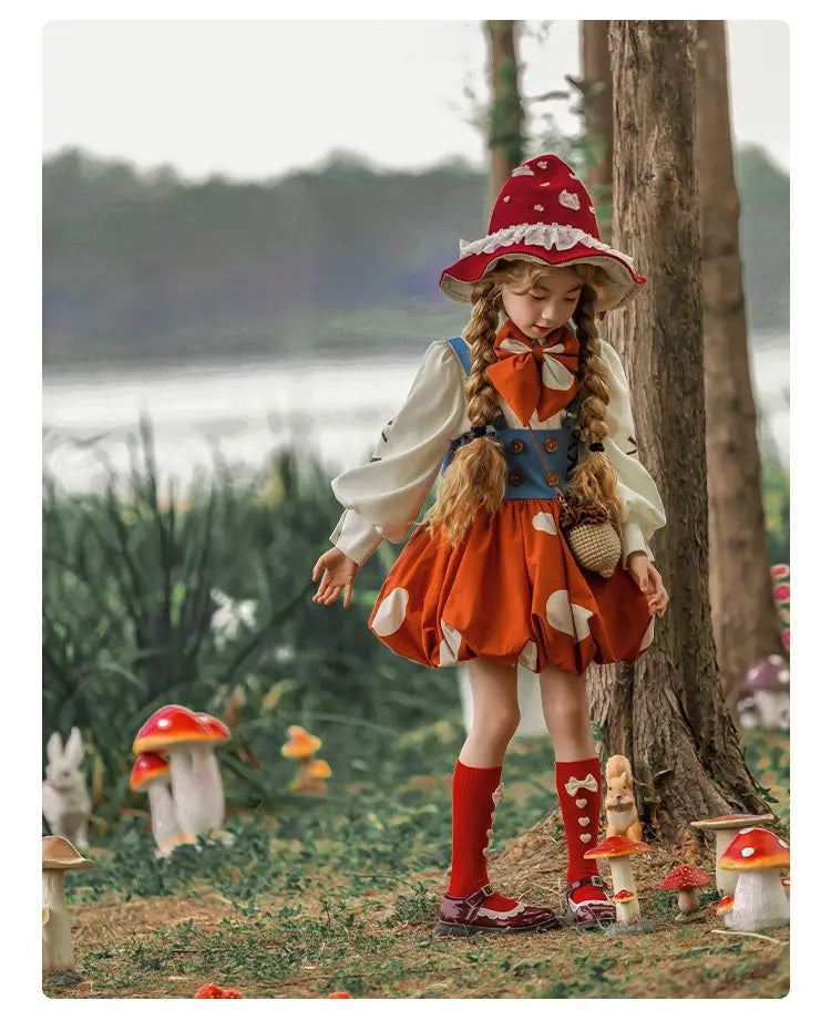 A young girl modeling a whimsical fairy tale costume in an outdoor forest setting with mushrooms and trees. The costume features a red and white polka-dot witch hat, a white long-sleeve blouse, a blue vest, an orange skirt with white polka dots, red knee-high socks, and red shoes.