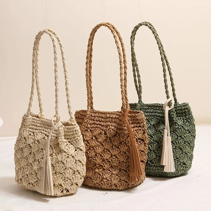 Three woven straw bucket bags in beige, tan, and sage green, each with braided shoulder straps and a tassel detail, displayed against a neutral cream background.